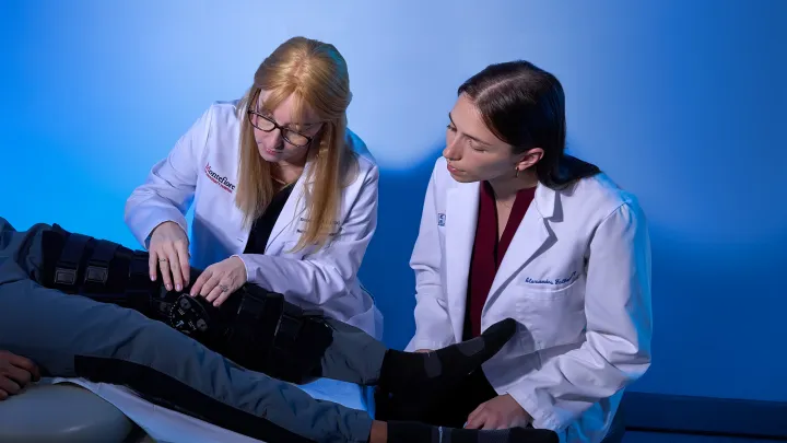 Two medical students in white coats examining a patient's leg brace in a clinical training setting.
