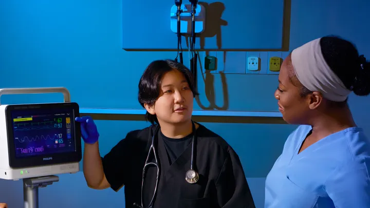 wo healthcare professionals in scrubs stand beside a patient monitor, with one pointing at the screen and the other listening, engaged in a clinical discussion in a hospital setting.