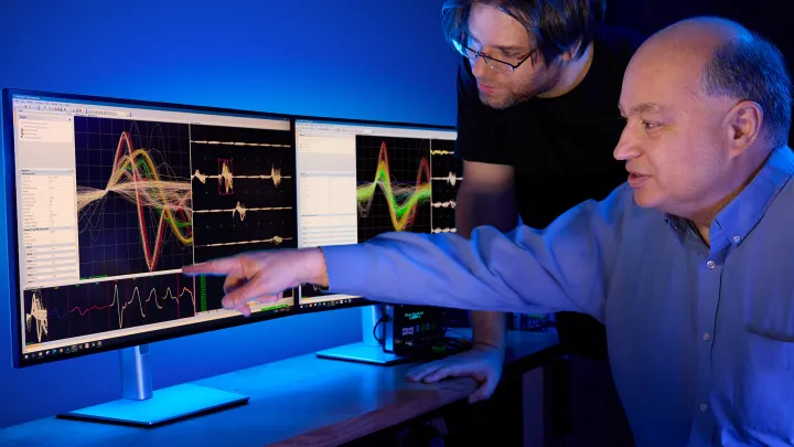 Middle-aged man and younger man analyzing colorful data visualizations on computer screens in a high-tech lab setting.