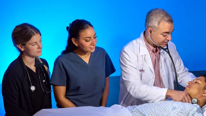 Male physician teaches medical students at a bedside exam with a patient in a clinical learning environment.