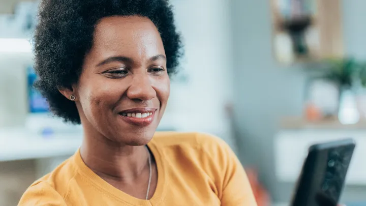 Woman in a yellow sweater smiling while looking at her smartphone at home.
