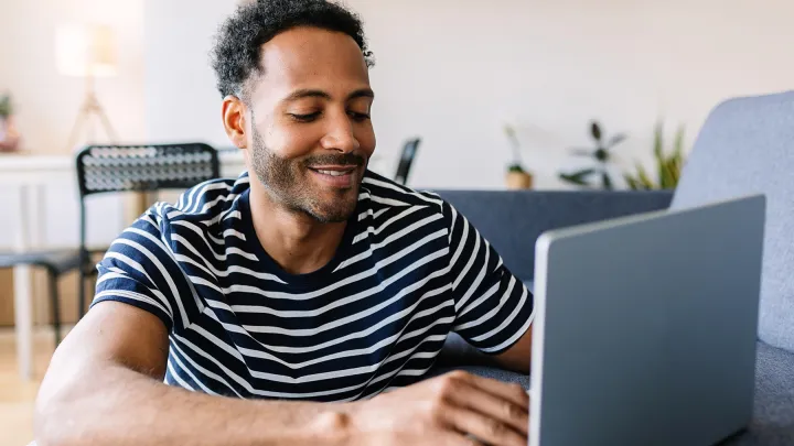 Young man in a striped shirt smiling while working on a laptop in a modern home environment.