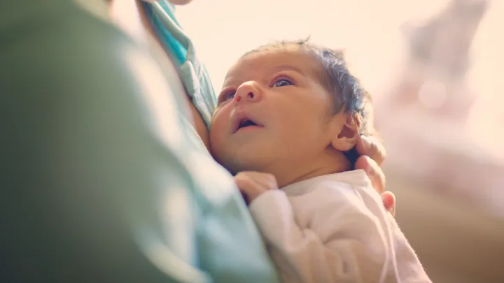 Newborn baby gazing up while being held gently in a parent's arms with soft natural light in the background.
