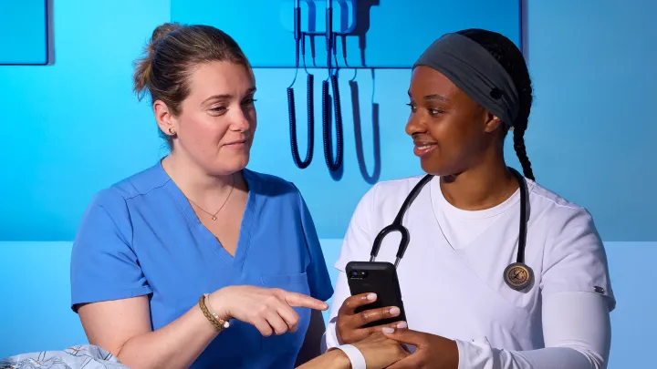 Two nurses review patient information on a mobile phone at the bedside while providing clinical care.