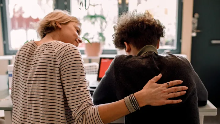 A supportive person places a hand on the back of a teen, offering comfort at home.