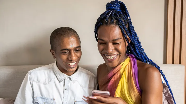 A non-binary person and transgender woman sit close together, smiling while looking at a smartphone indoors.