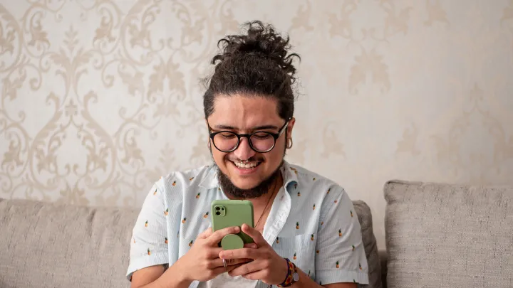 A transgender man wearing glasses and a patterned shirt smiles while using a smartphone on the couch.