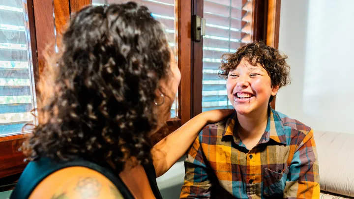 A parent and a transgender teen share a joyful moment together, smiling warmly indoors.