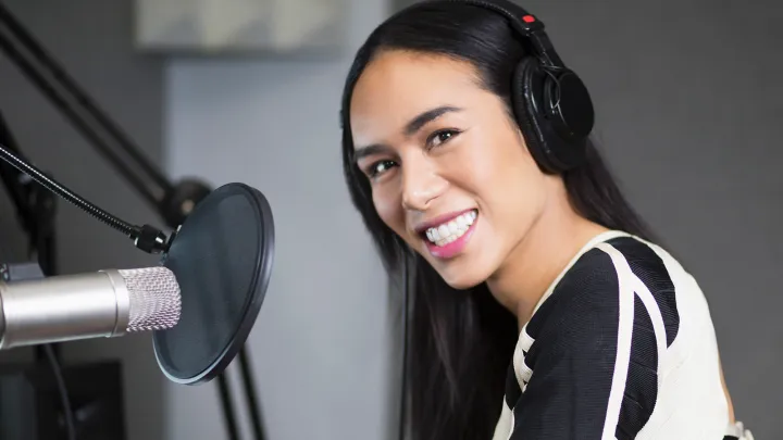 A transgender woman in headphones smiles while speaking into a studio microphone, recording a podcast.