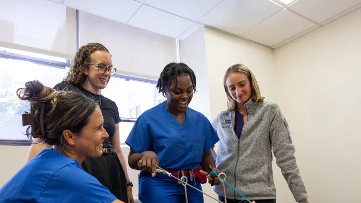 A group of four people, including medical trainees in scrubs, practice laparoscopic skills on a training model while smiling and collaborating.