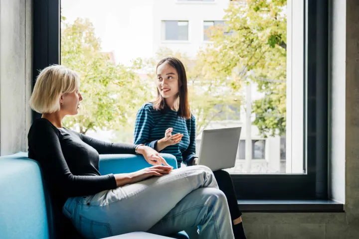 Two women talking, seating with a window view of some trees