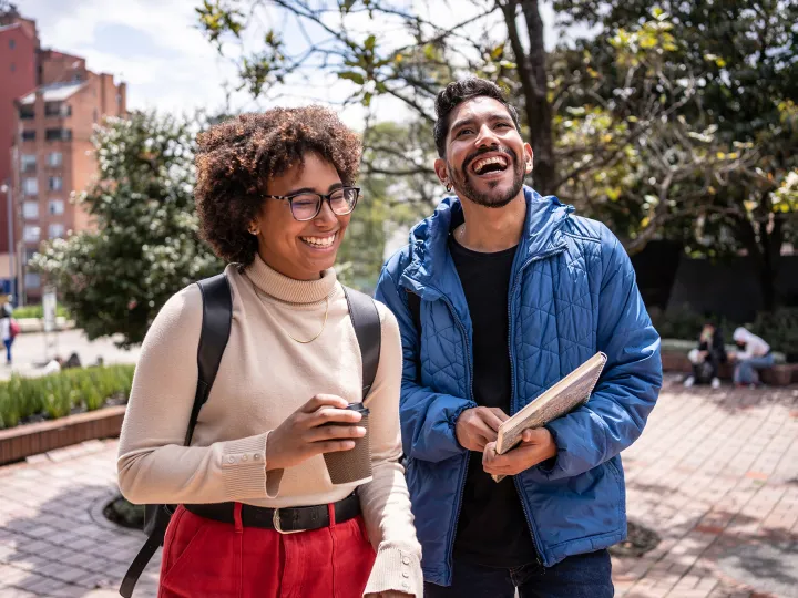 Two students standing outside on campus, smiling and laughing while holding coffee and notebooks.