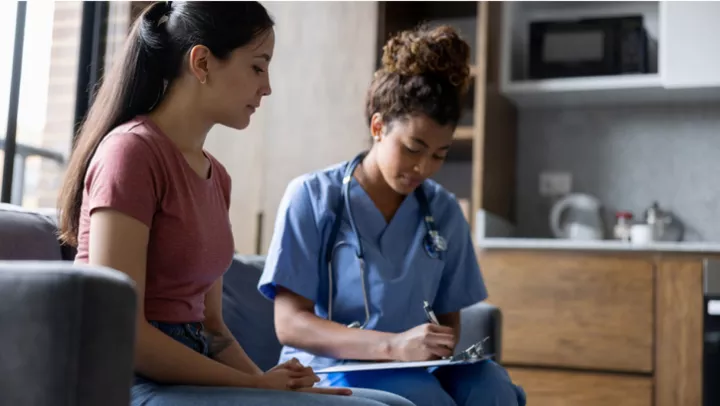 A long-haired woman in jeans and a coral top talks to a healthcare provider as they fill out a document.