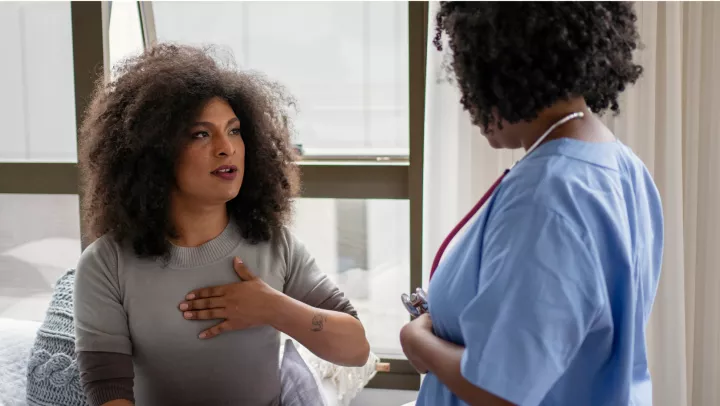 A black woman wearing a gray sweater sits near a window and speaks to a black female healthcare worker wearing blue scrubs.