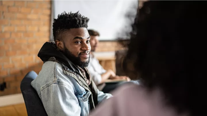 A black man with a beard and a fade hairstyle sits in a chair, listening to a blurry, indistinct speaker sitting to his right.