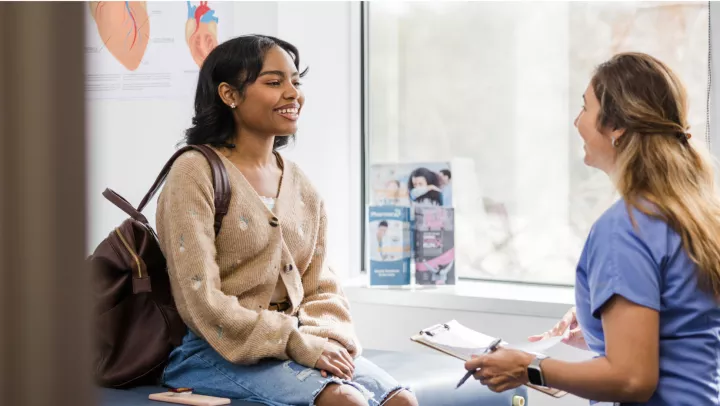 A black teenage girl wearing jeans and a beige cardigan sits in a healthcare clinic and speaks with a blonde nurse.
