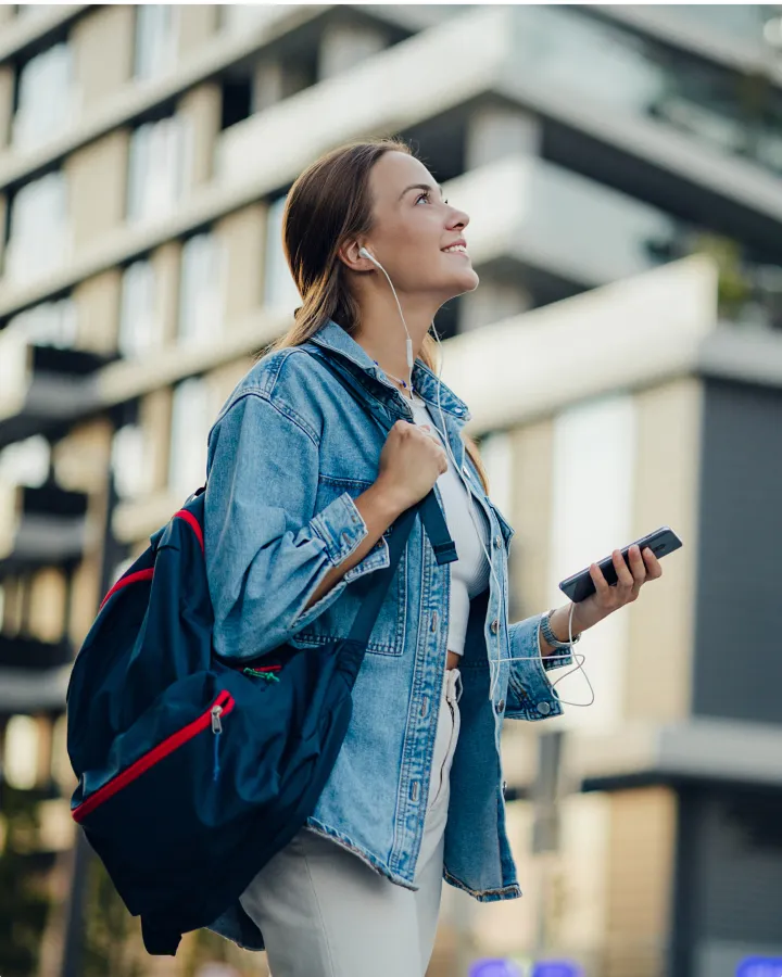 Young woman with a backpack and earphones walks through a city street, smiling and looking up while holding a phone.