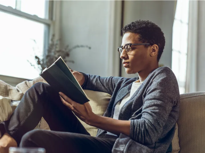 Young man with glasses sits on a sofa at home, reading a book by a sunlit window.