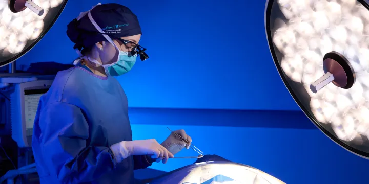 Surgeon in blue scrubs and mask preparing tools under surgical lights.
