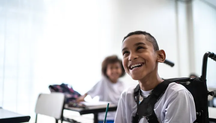 Smiling boy in a wheelchair wearing a white shirt, sitting in a classroom with another child blurred in the background.