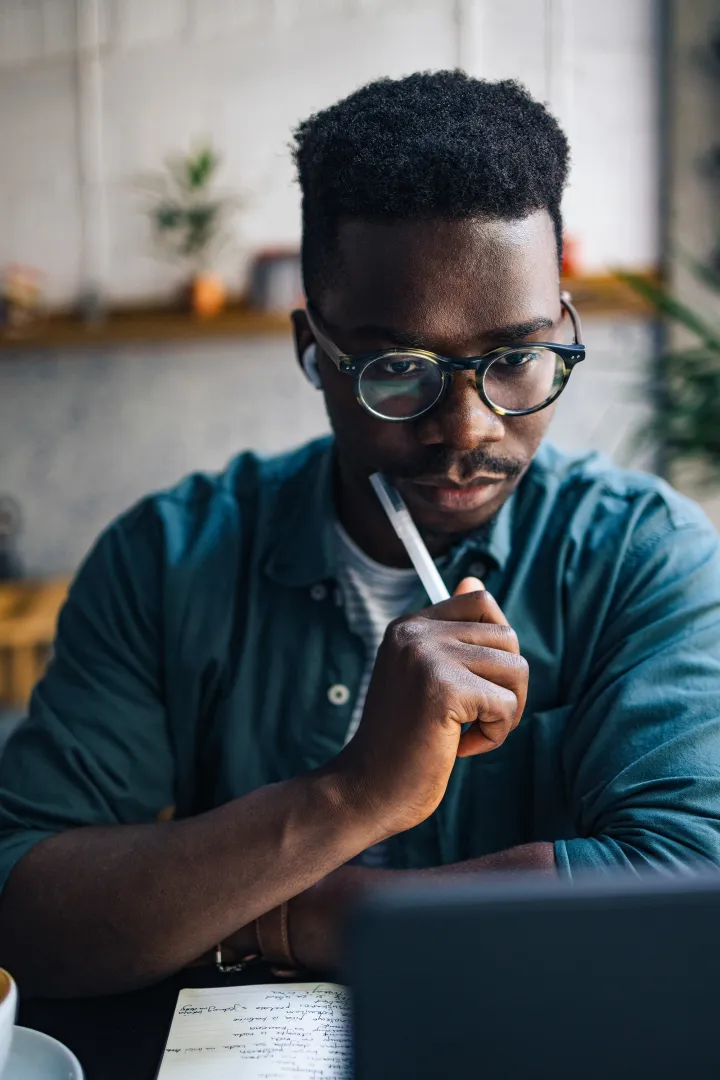 Focused man wearing glasses and earphones studies at a table with a notebook and laptop in a cozy café.