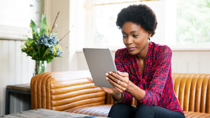 A woman holding an iPad sits on a couch in a bright room.