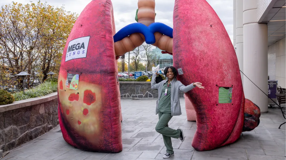 A smiling Montefiore Einstein staff member in green scrubs poses between two giant inflatable lung models at the MEGA Lungs interactive exhibit, part of the annual Lung Cancer Screening community event.
